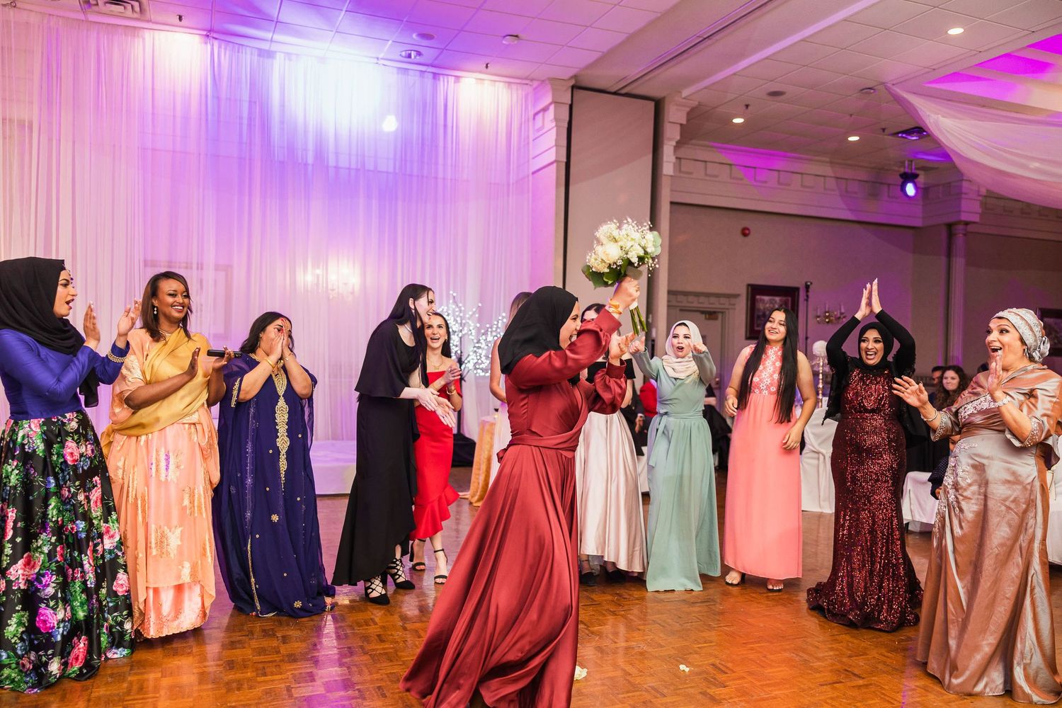 Wedding guests in colorful formal attire dancing in a line at a reception venue with white drapery.
