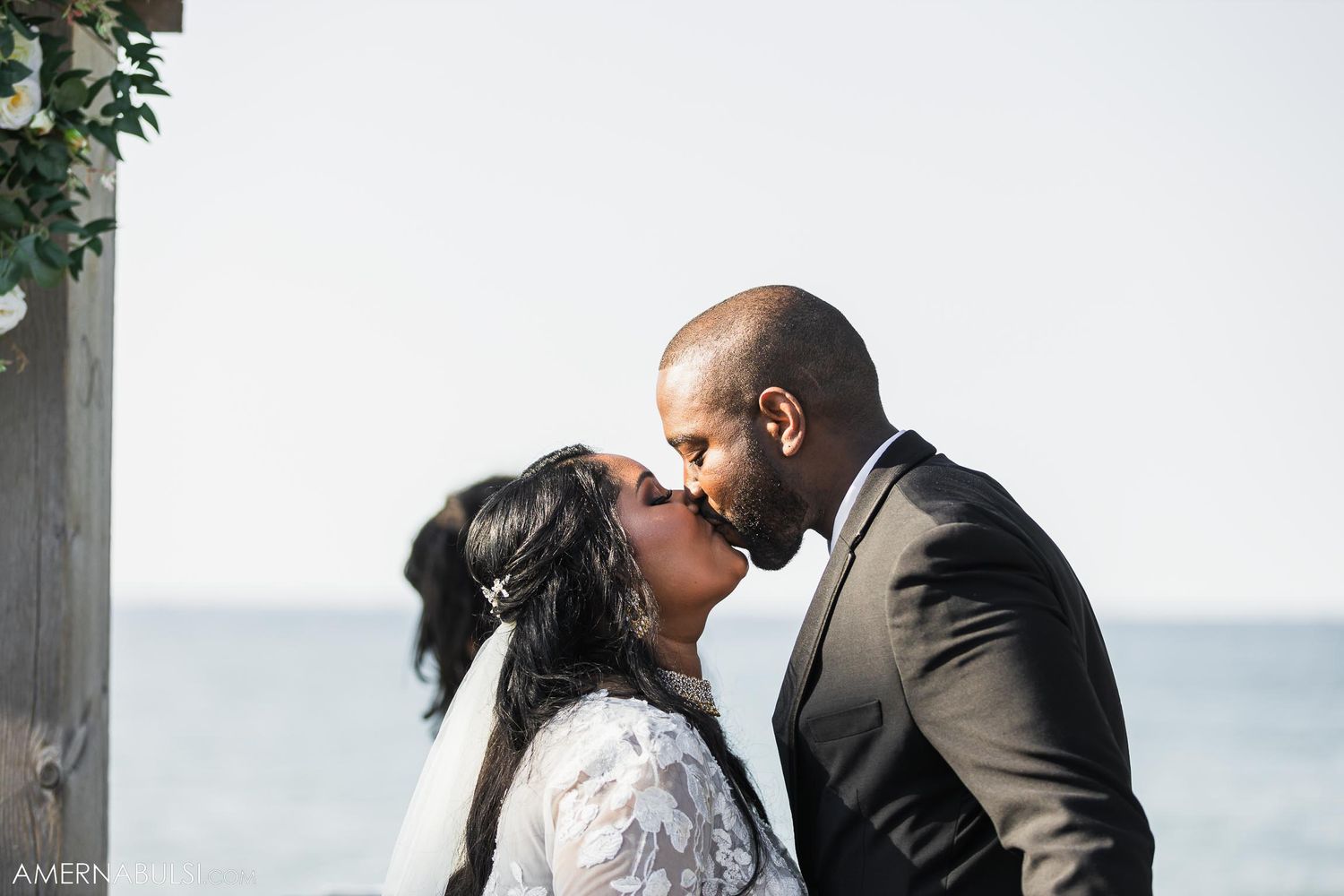 Intimate wedding kiss under floral-decorated pergola with ocean backdrop.