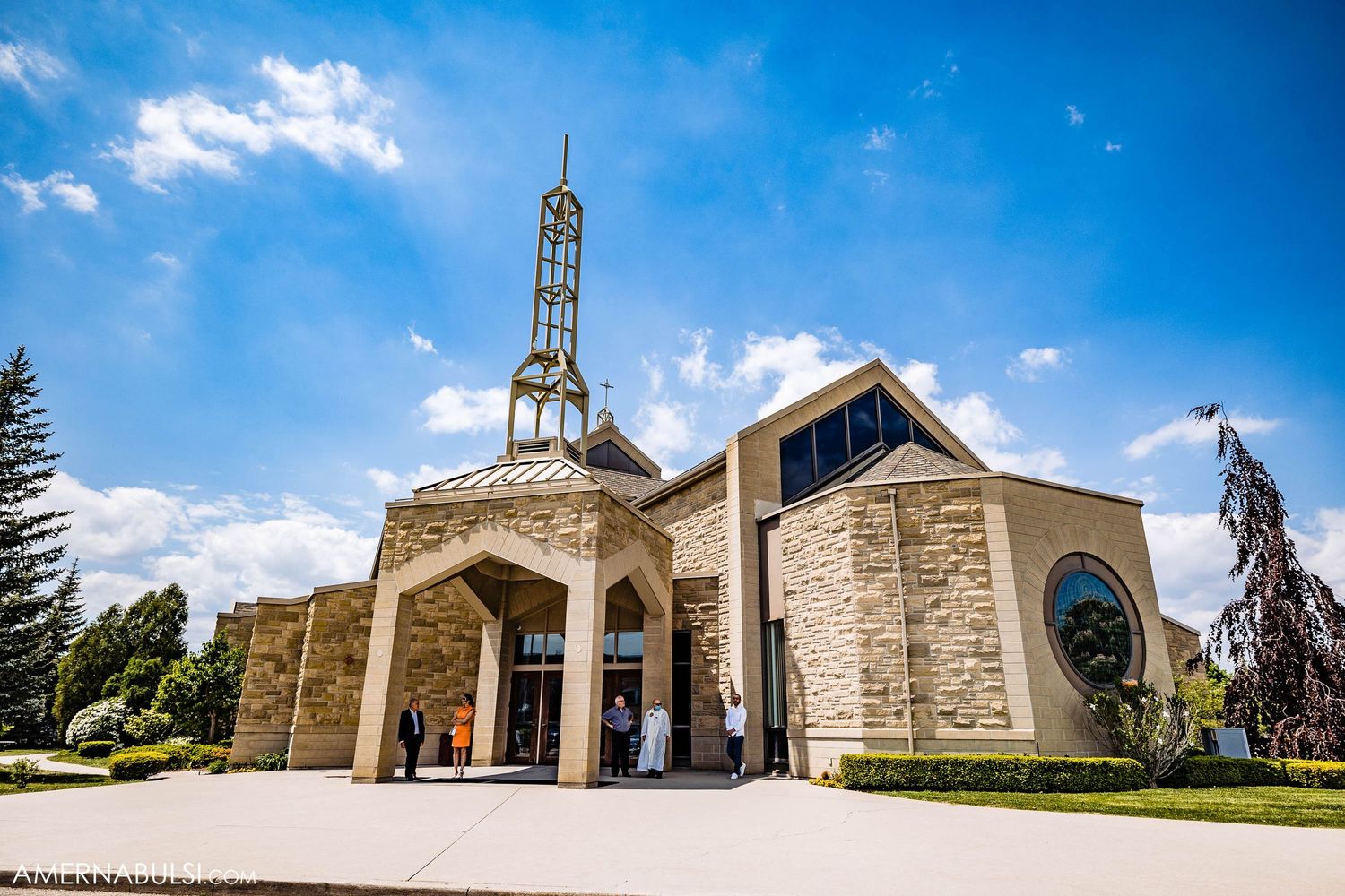Modern stone church exterior with tall spire against bright blue sky.