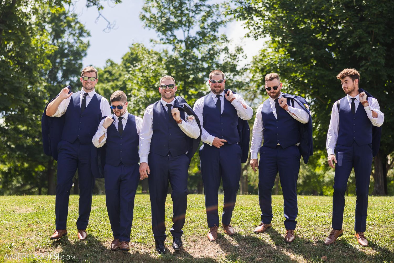 Groomsmen in navy vests and ties pose playfully outdoors on sunny day.