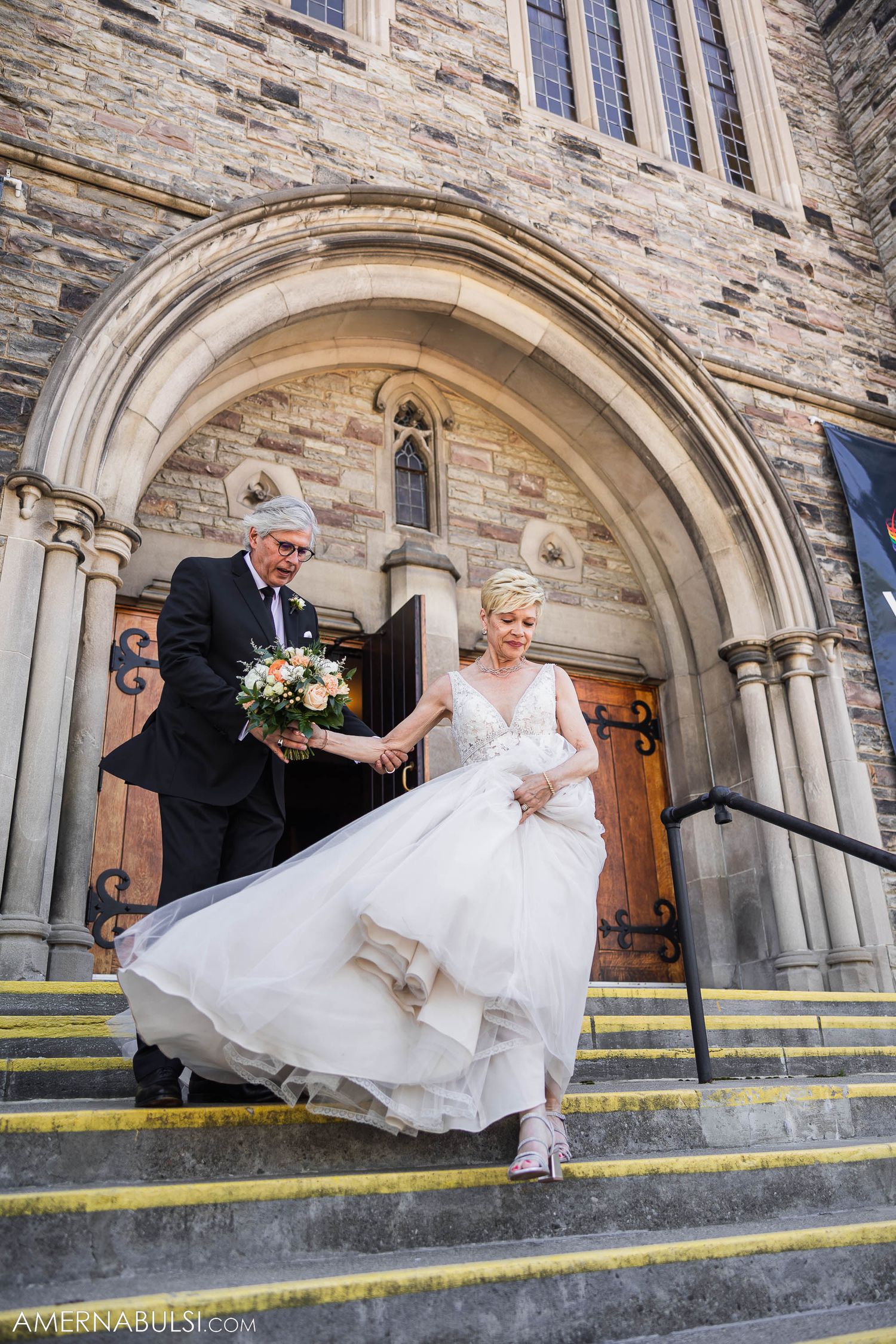 A couple descends stone church steps while holding a floral bouquet.