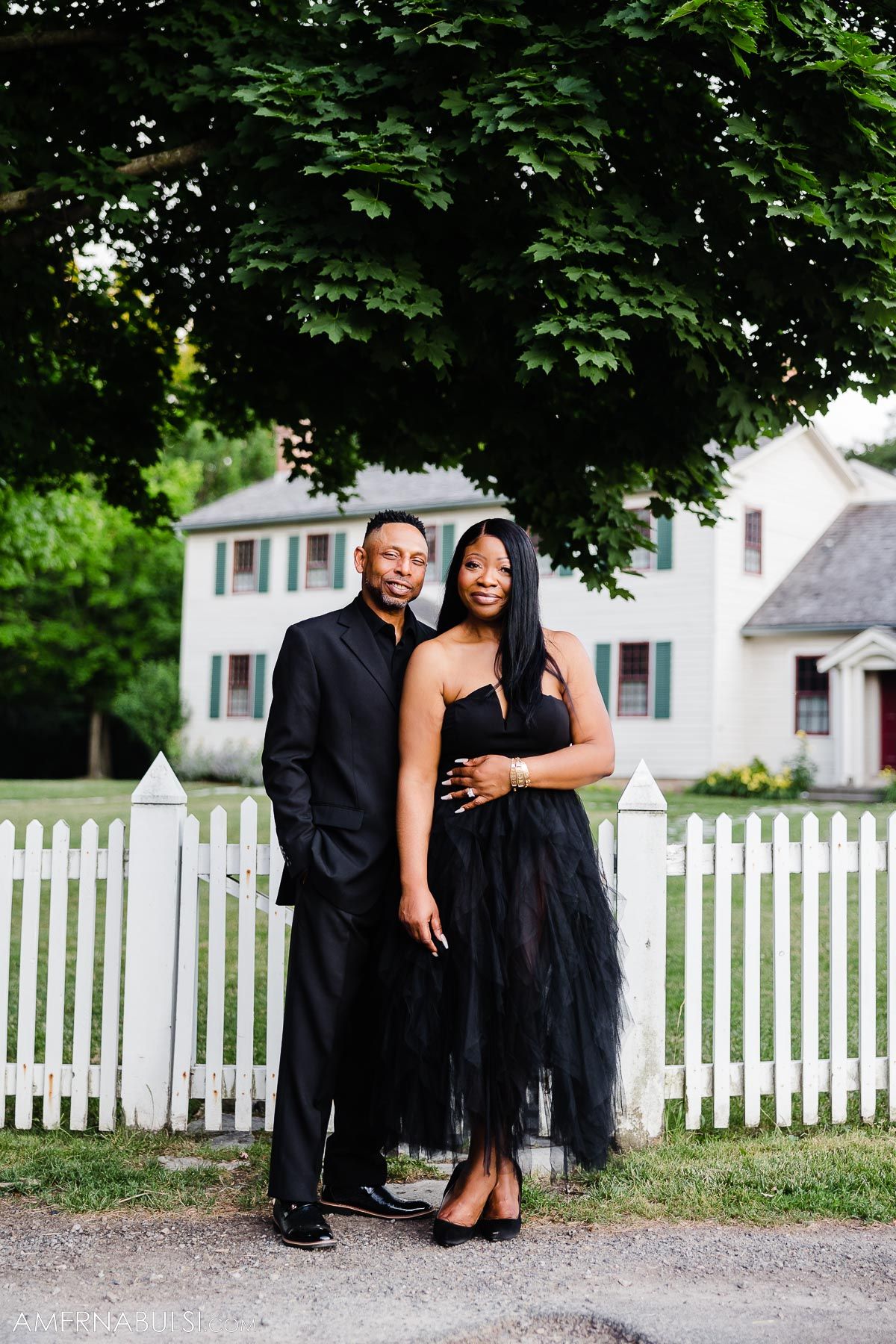 Elegant couple in black formal wear pose in front of white picket fence and colonial house.