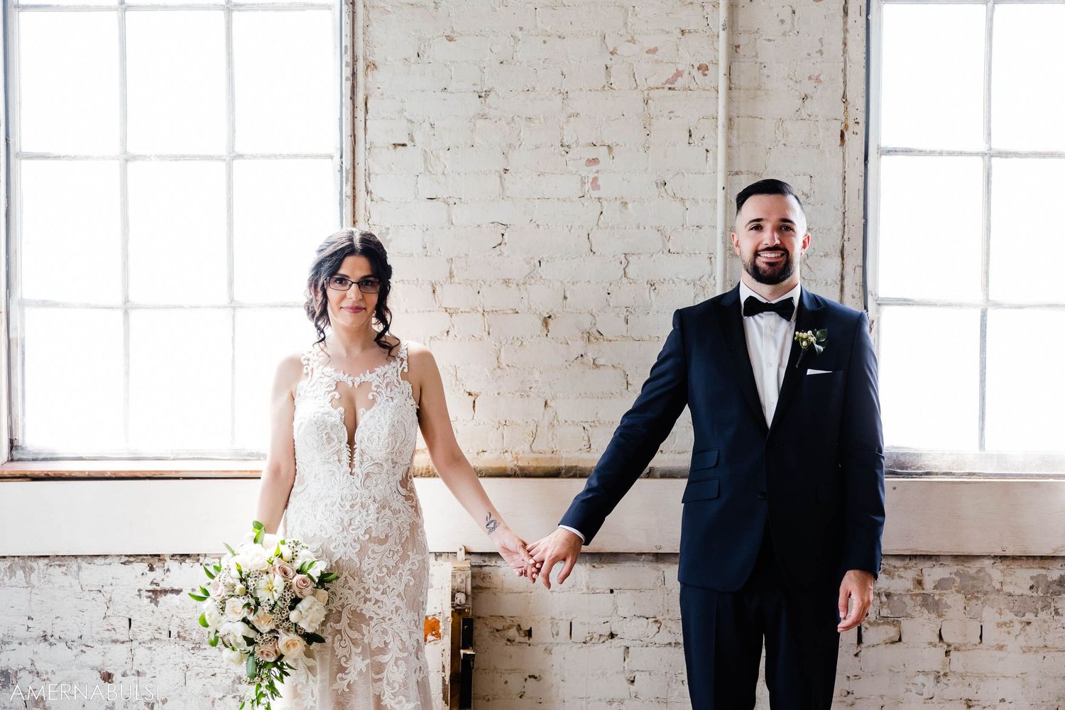 A bride in a lace gown and groom in a navy suit pose together against a white brick wall with large windows.