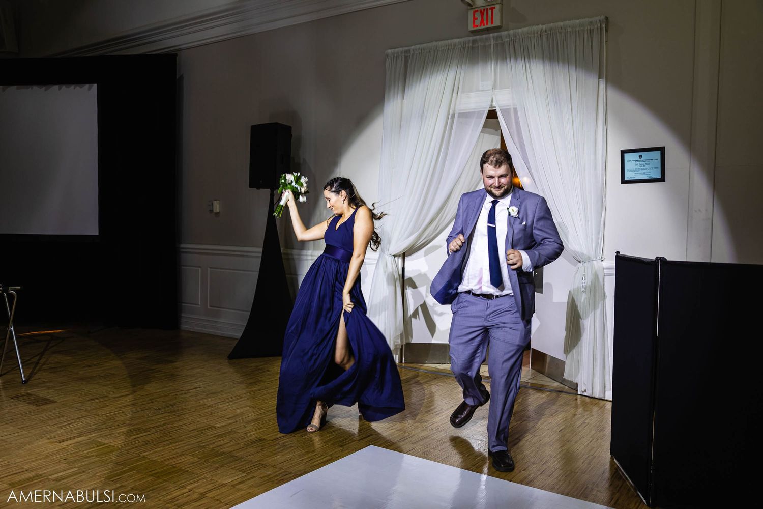 Wedding party members in navy blue and grey attire dance joyfully across a reception dance floor.