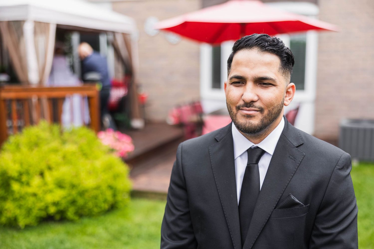 A well-dressed man in a black suit smiling outdoors with greenery in the background.