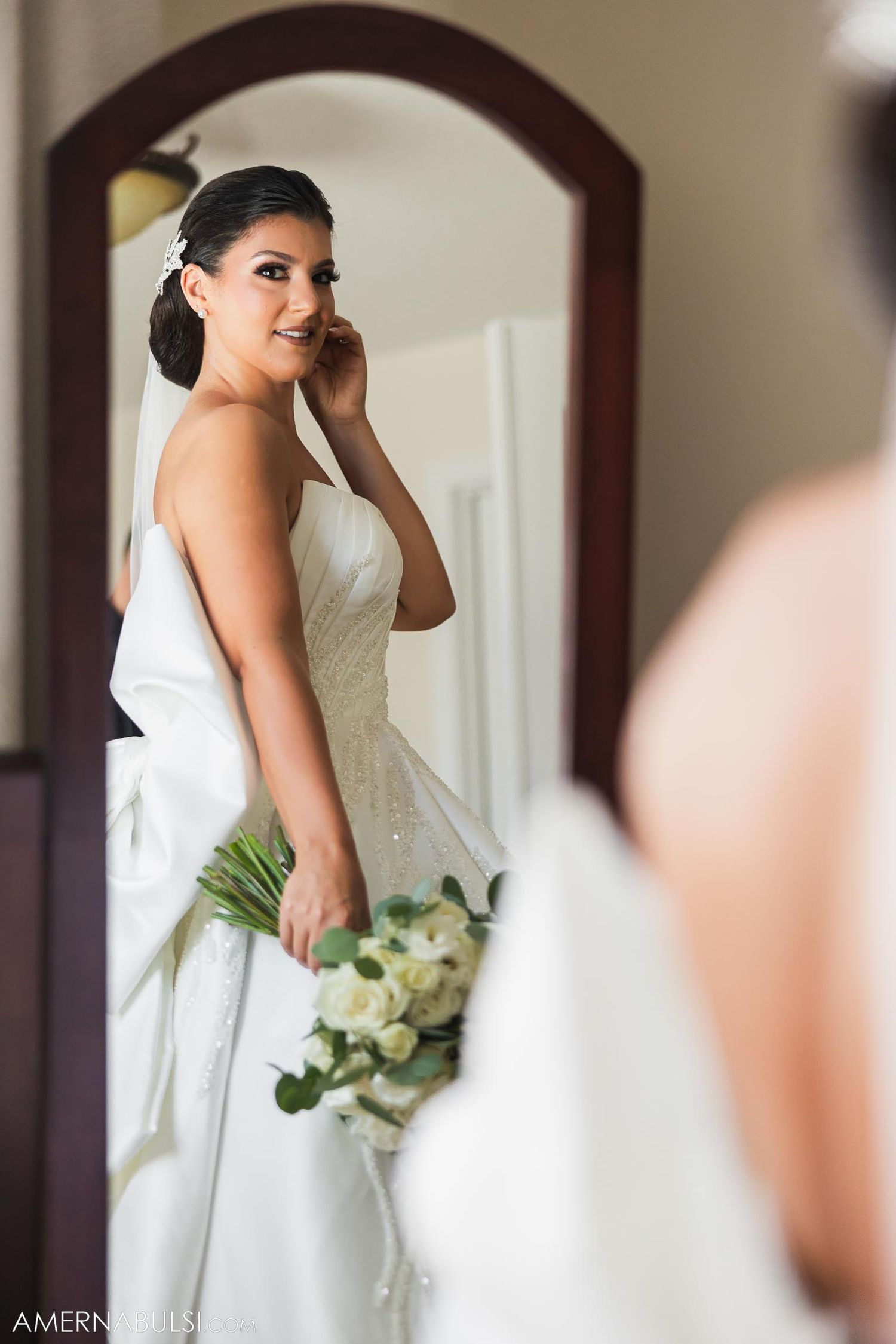 Bride wearing strapless dress looks in mirror while holding white flower bouquet.