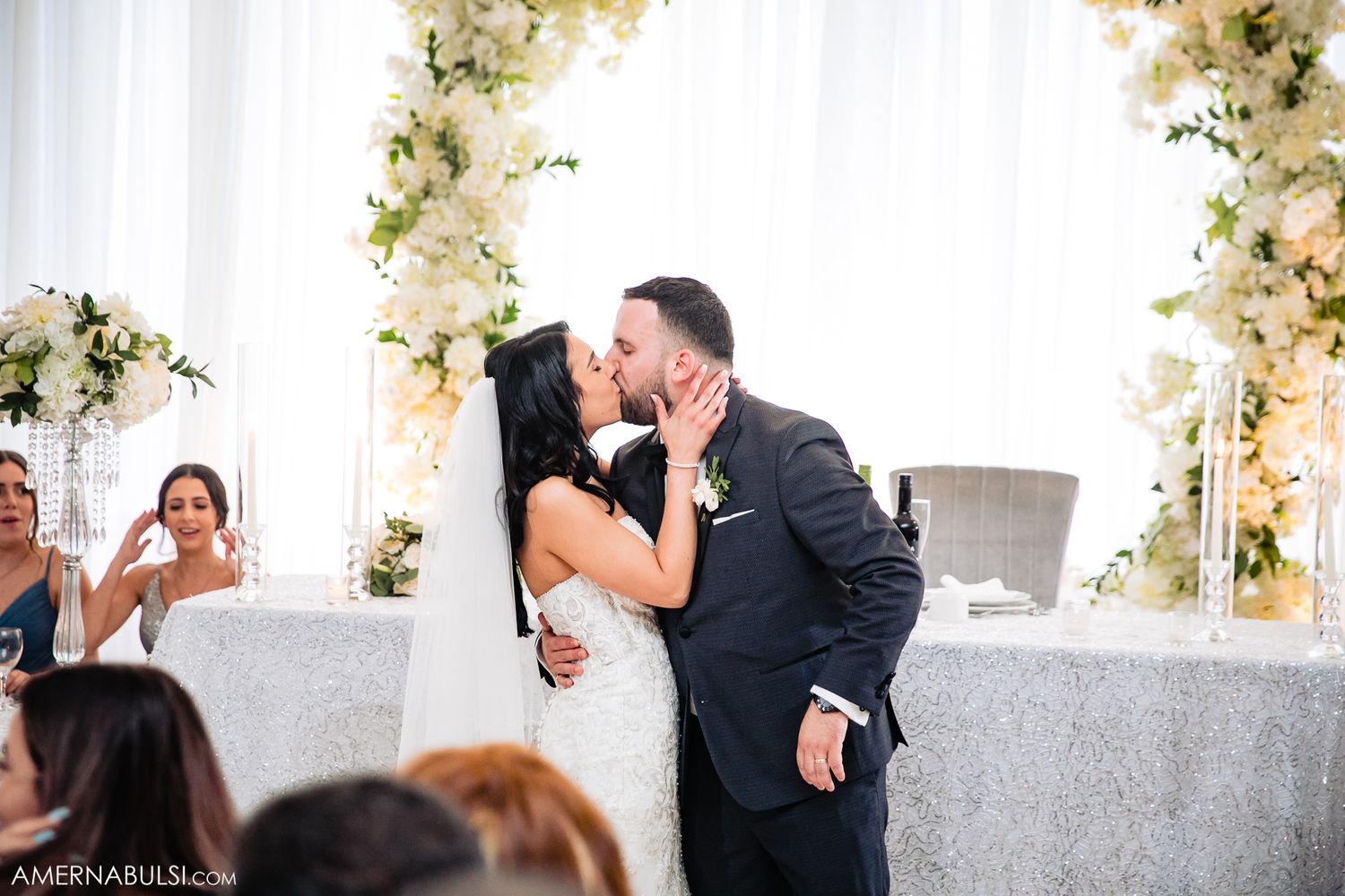 Couple sharing romantic moment under decorative white floral arch at wedding ceremony.