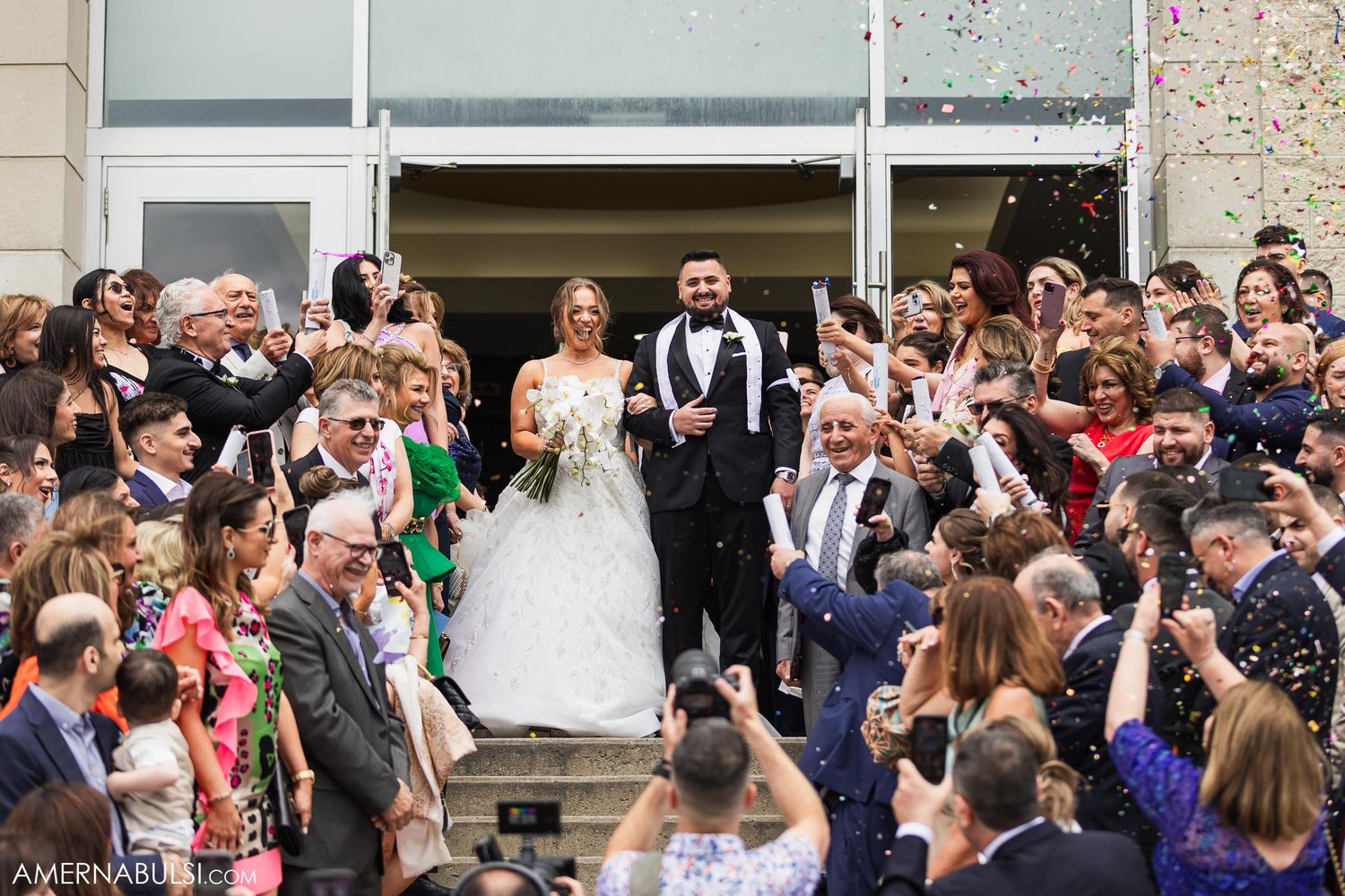 Wedding couple exits building to celebration with crowd throwing confetti amid joyful atmosphere.