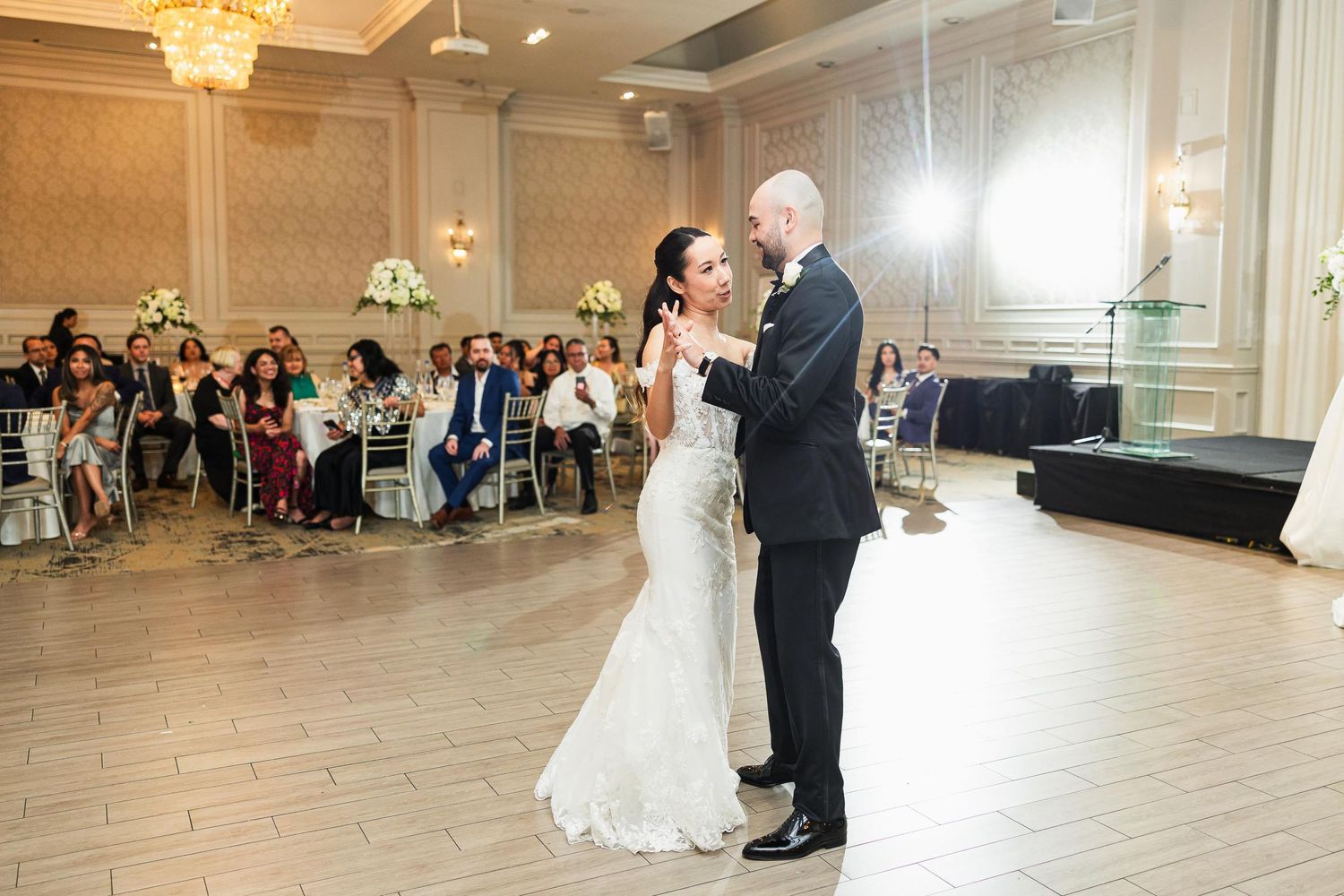 A couple performs their first dance in an elegant ballroom with guests watching from decorated tables.