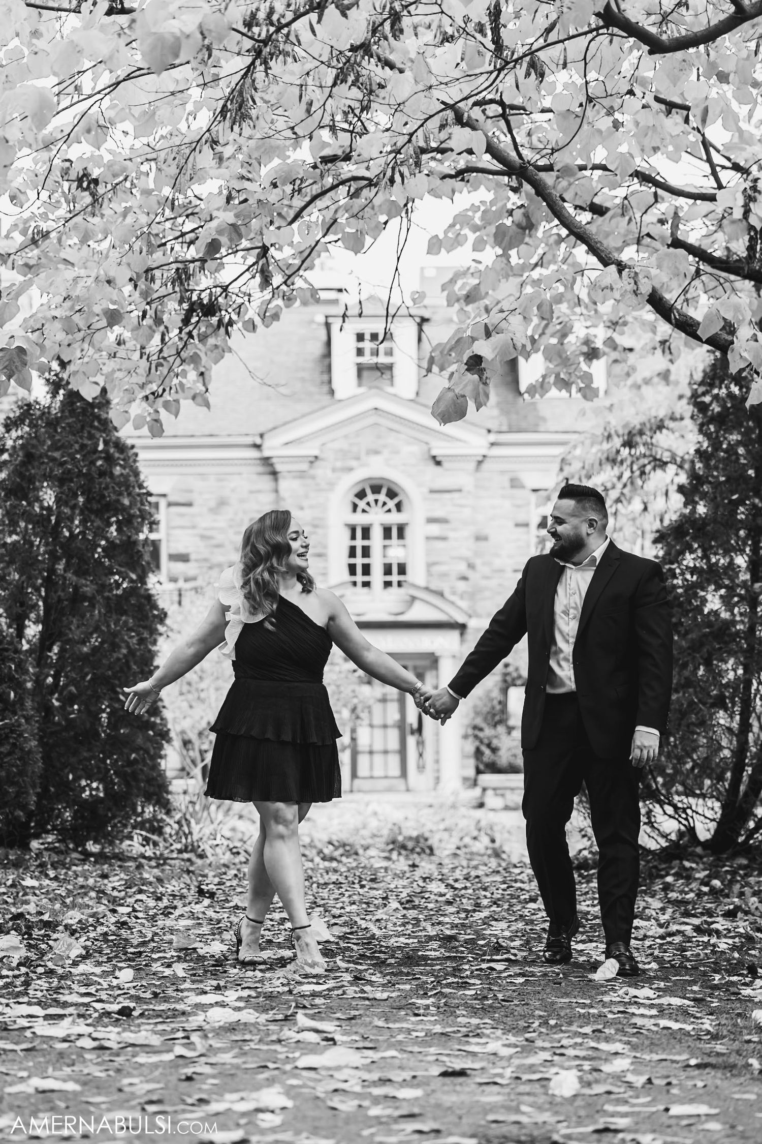 A couple holds hands while walking near a historic white building with autumn leaves overhead.