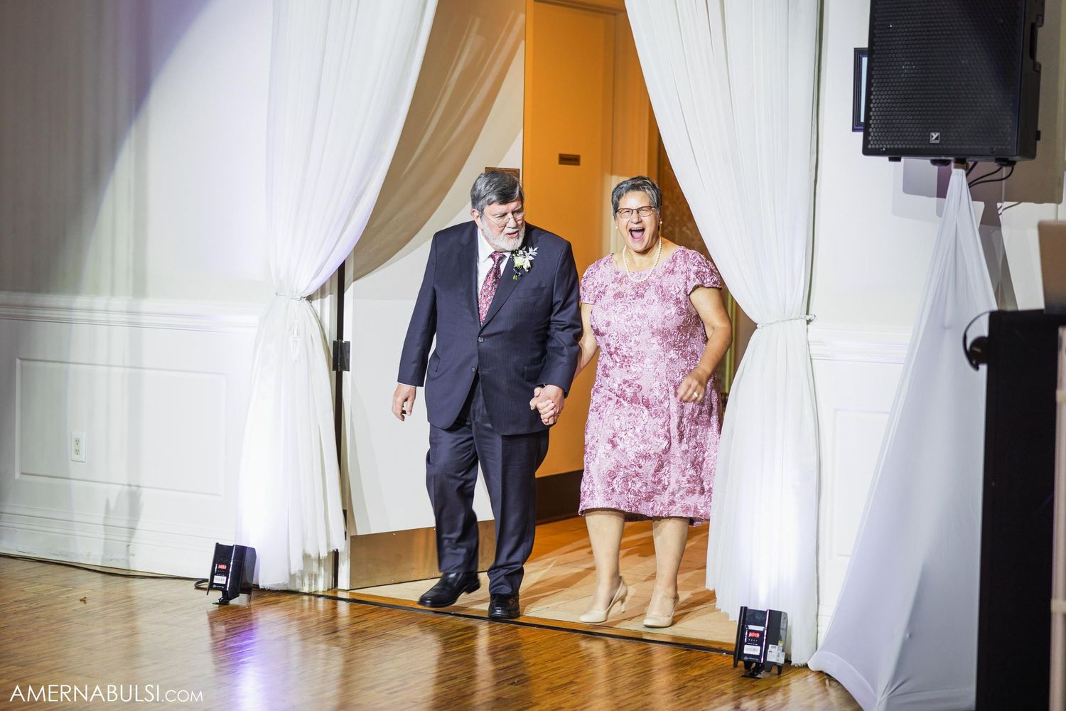 Couple in formal attire make dramatic entrance through white draped doorway at reception.