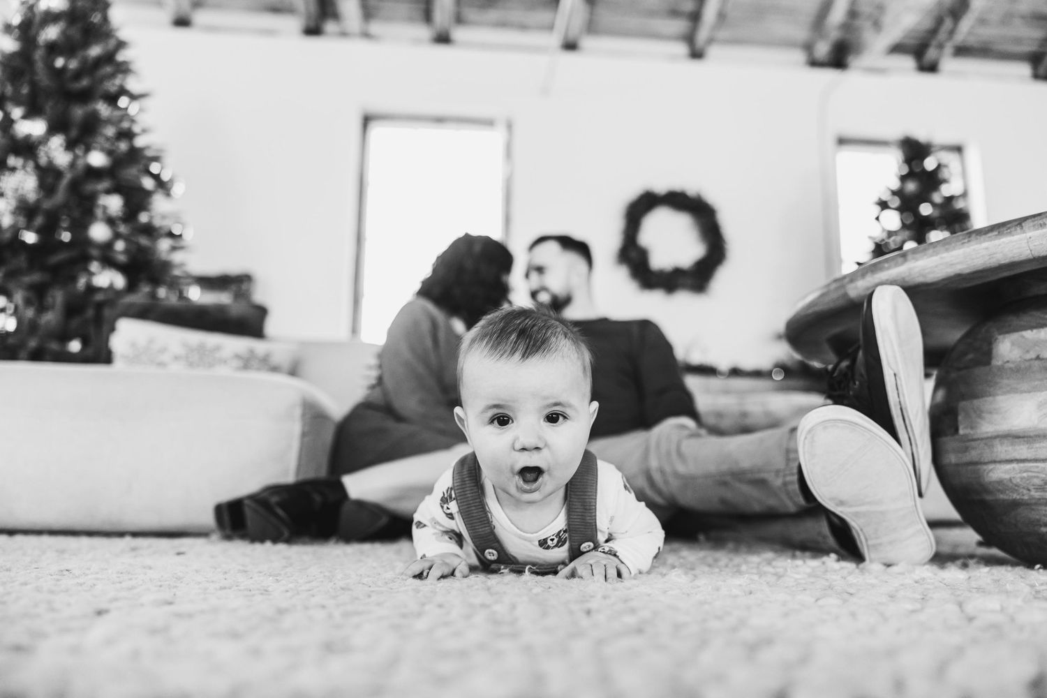 Black and white photo of baby crawling while parents relax on couch in festively decorated room.