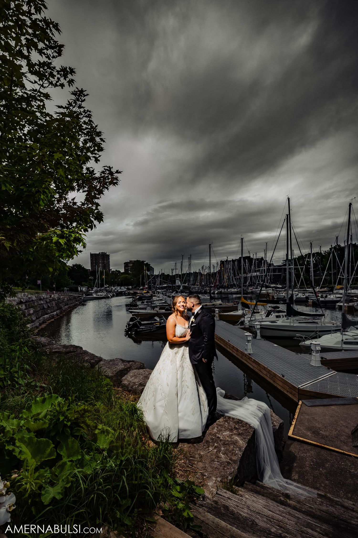 Dramatic wedding photo at marina with stormy sky and sailboats in background.