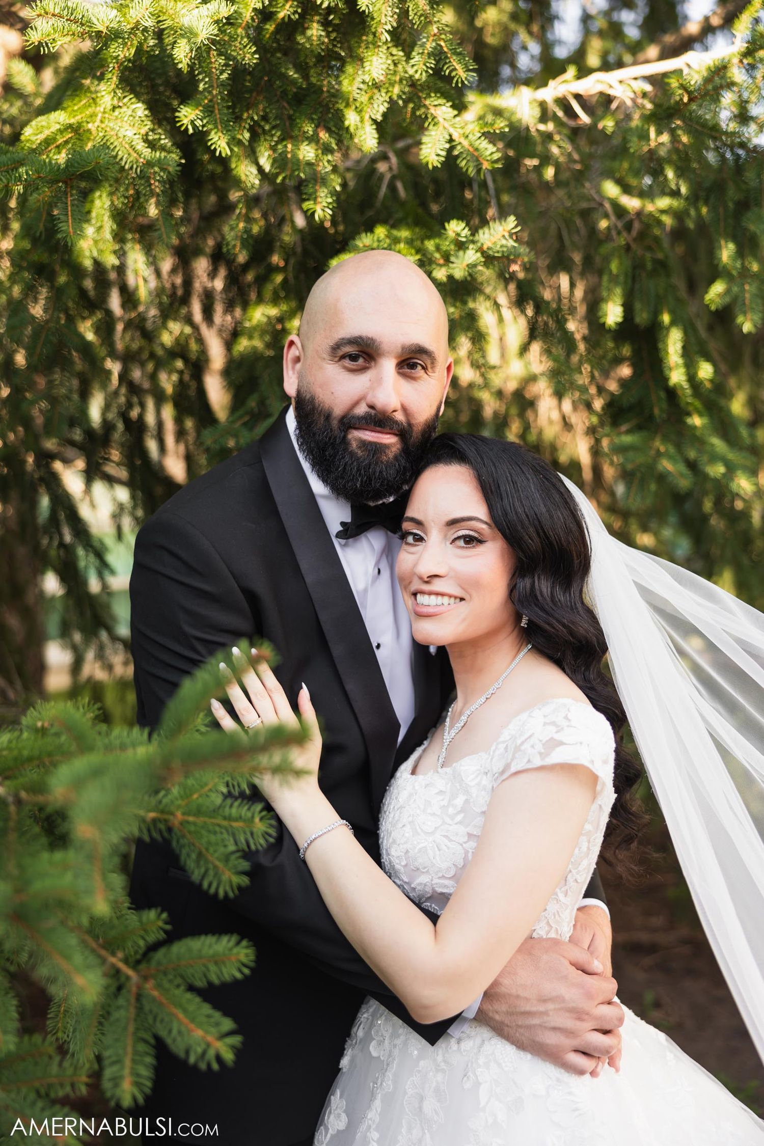 A couple poses together under evergreen branches during their wedding photoshoot outdoors.