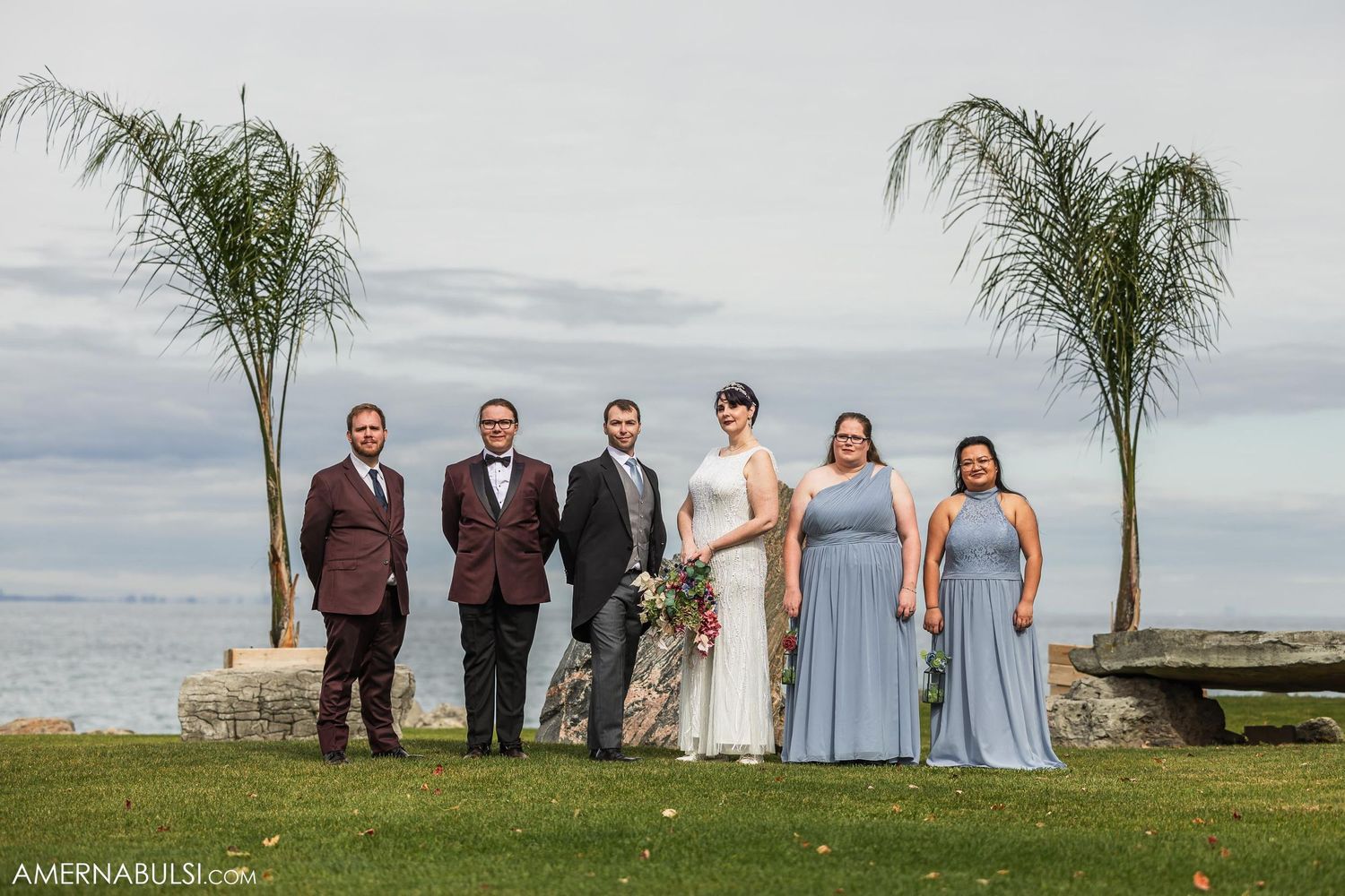 Wedding party poses on oceanfront lawn with tropical palm trees framing the scene.