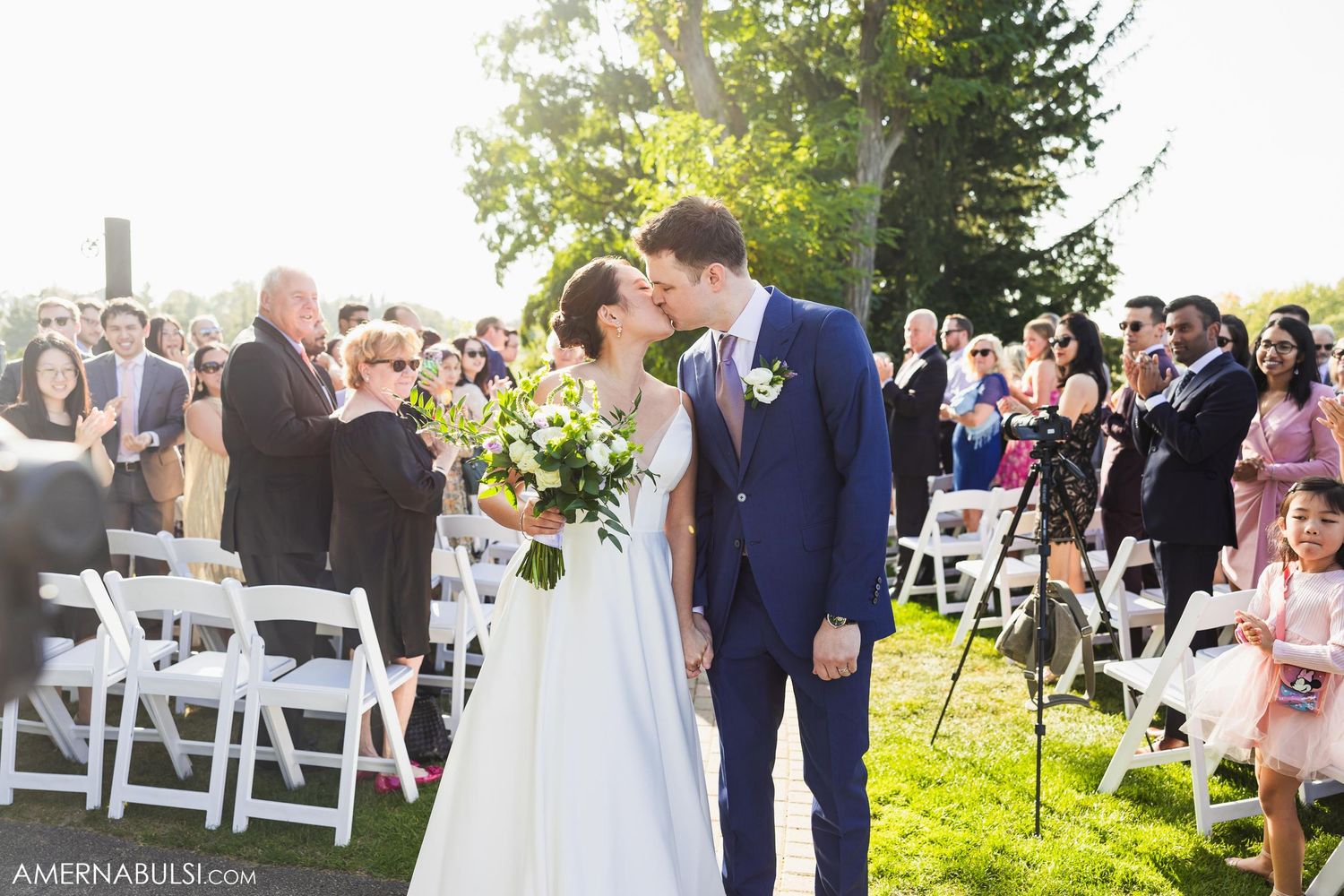 Newlyweds share first kiss during outdoor wedding ceremony surrounded by guests.