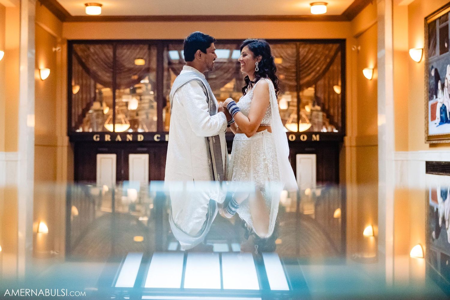 Couple in white Indian wedding attire dance in elegant room with mirrored floor.