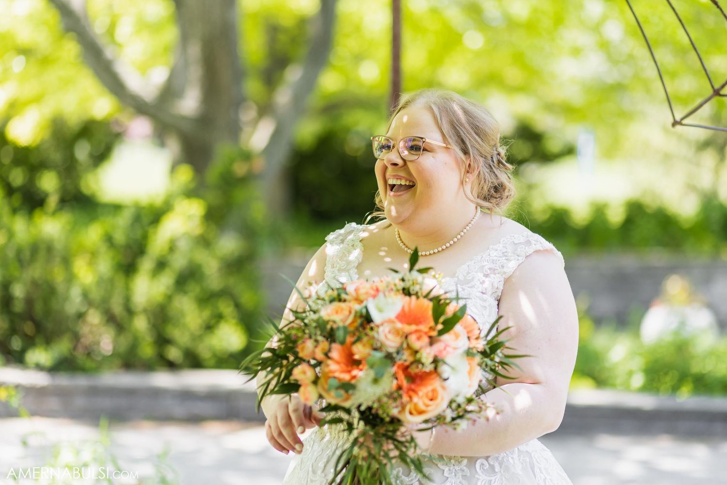 Bride laughs joyfully while holding beautiful peach and orange floral bouquet in outdoor garden setting.