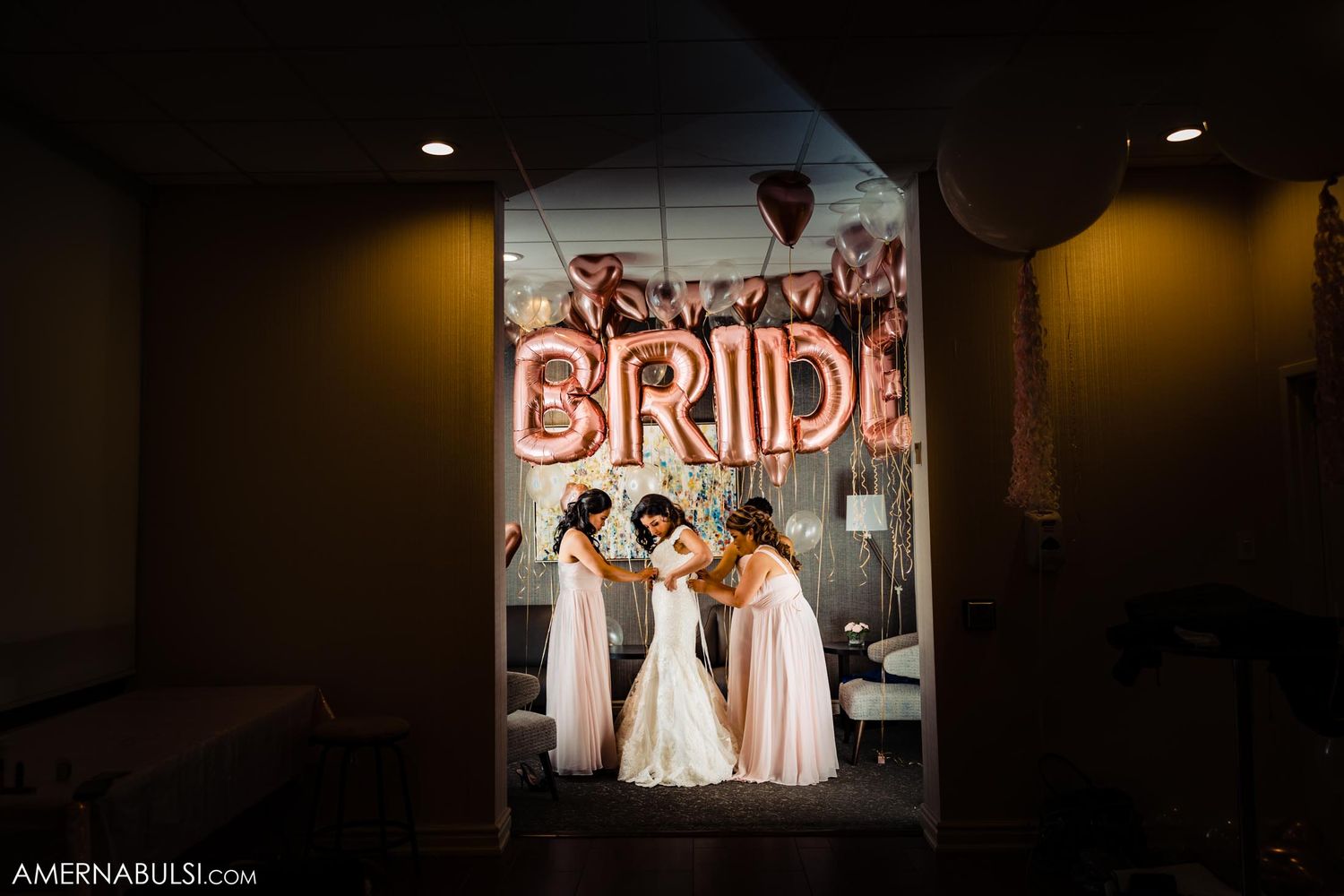 Pink BRIDE balloon letters illuminate a dark hallway where a bridal party gathers.