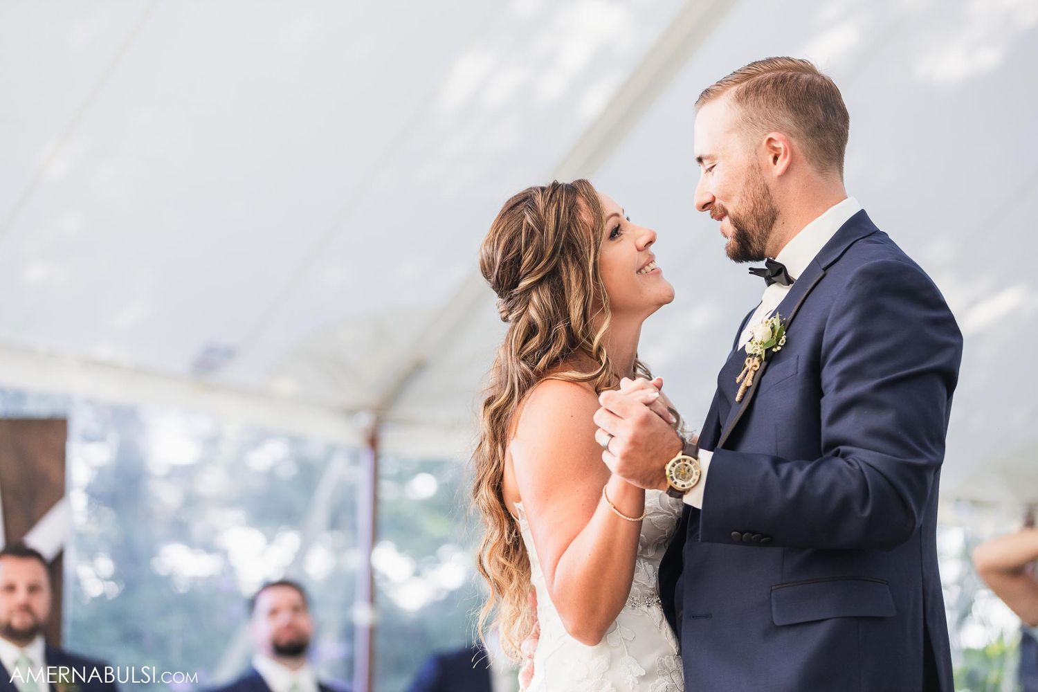Wedding reception under white tent with guests mingling around decorated tables and chairs.