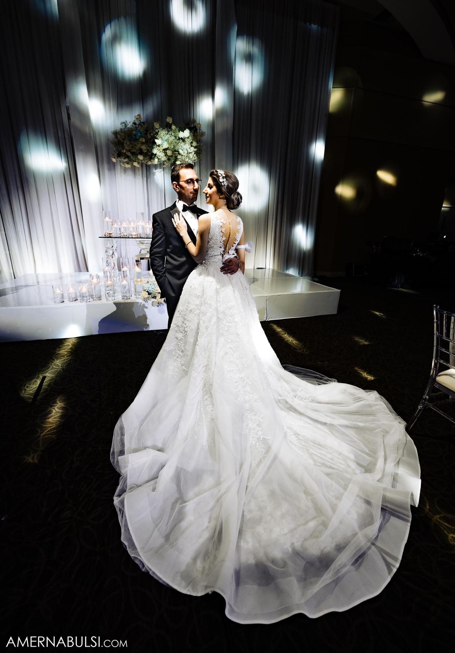 Elegant wedding couple in dramatic lighting with bride wearing flowing train dress and spotlight effects.