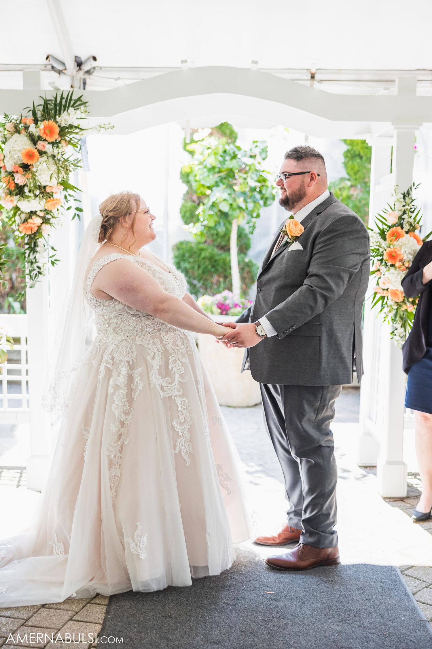 Couple holding hands during wedding ceremony under decorated white pergola.