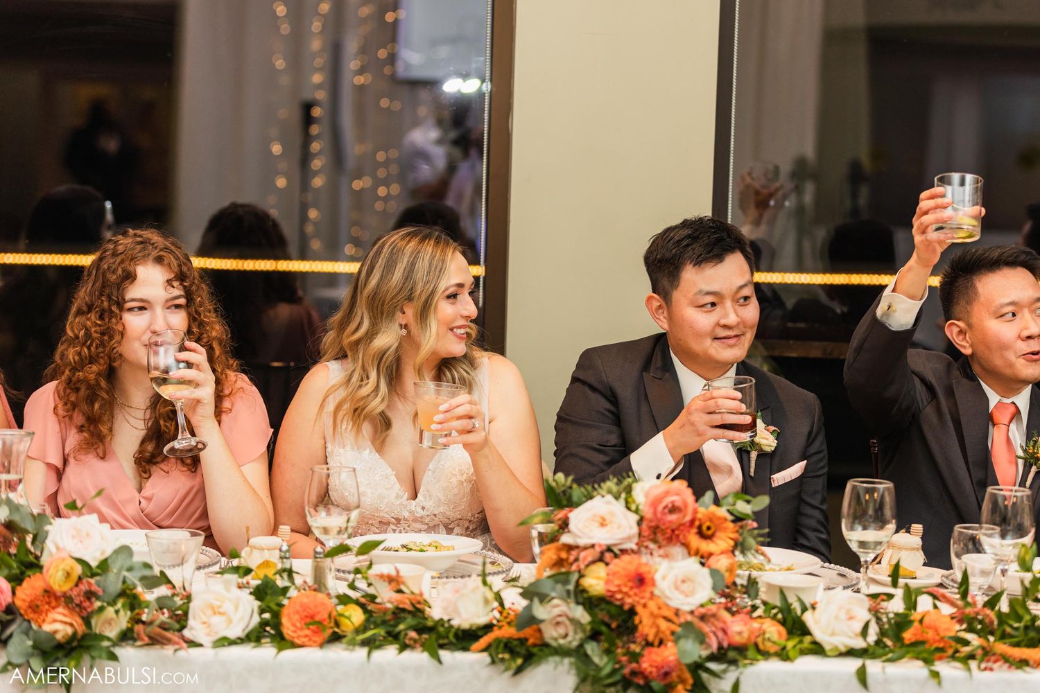 Wedding party members raise their glasses in a toast at an elegantly decorated reception table.