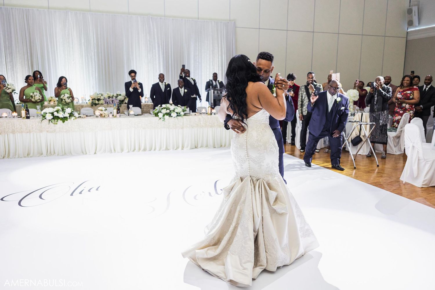 Couple sharing their first dance on white dance floor with wedding guests watching in background.