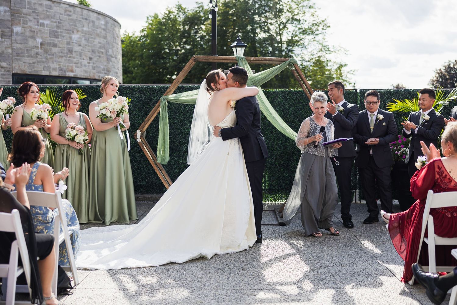 A bride and groom share their first kiss at an outdoor wedding ceremony with bridesmaids in sage green gowns.