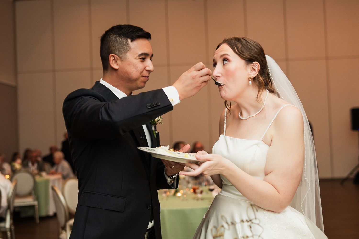 Groom feeding wedding cake to bride during reception celebration in elegant ballroom venue.