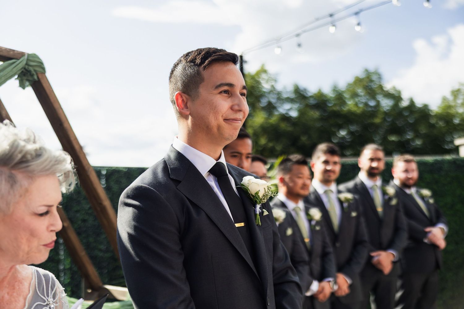 Groom smiling at the altar during outdoor wedding ceremony with groomsmen standing in line behind him.