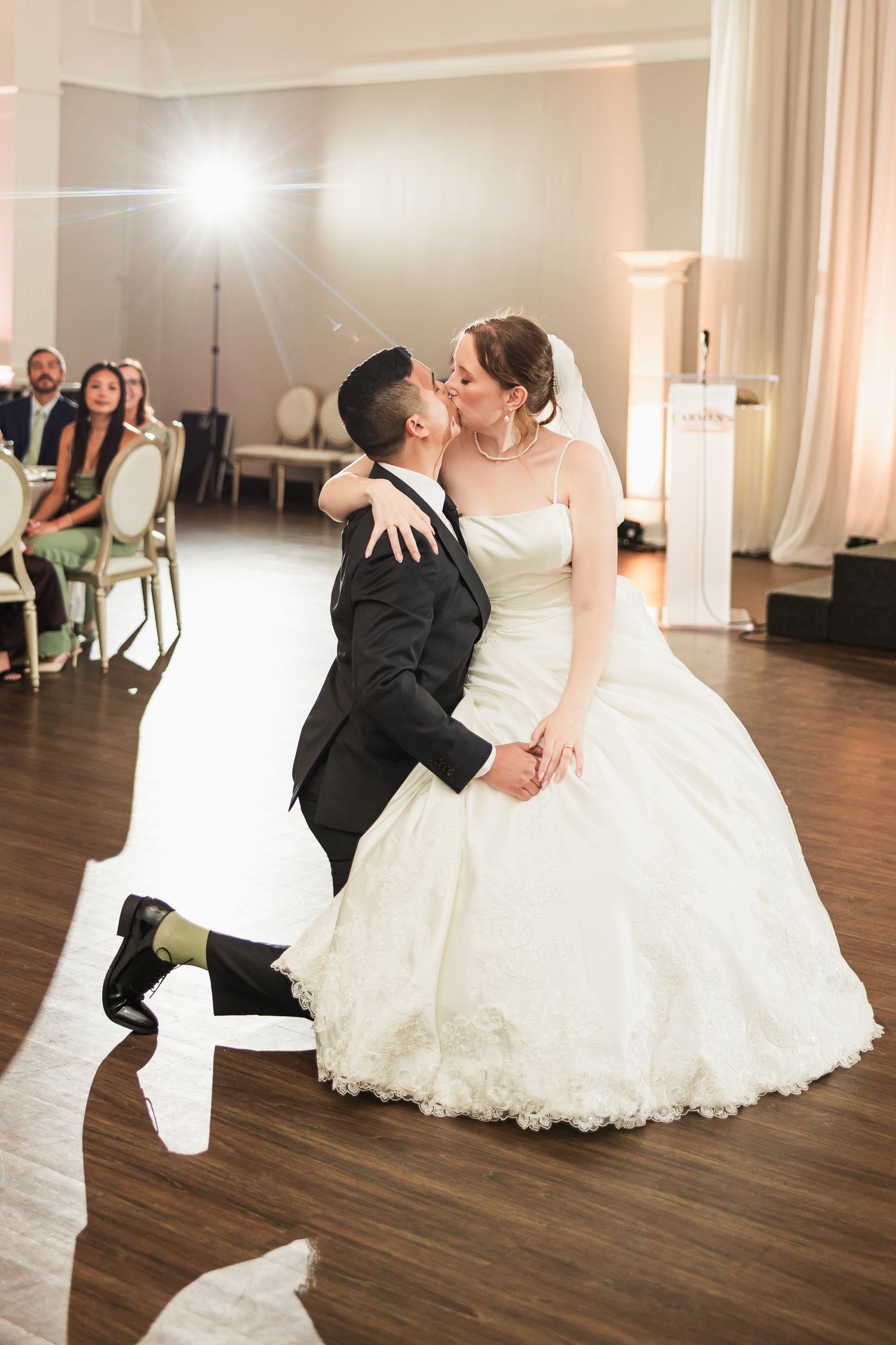 Groom kneels on dance floor kissing bride in a white ballgown during wedding first dance.