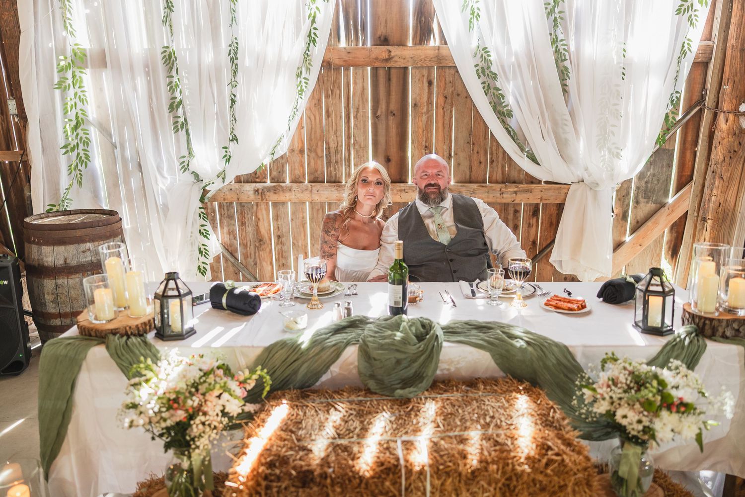 Bride and groom seated at rustic barn wedding sweetheart table with candles, greenery, and white draping.