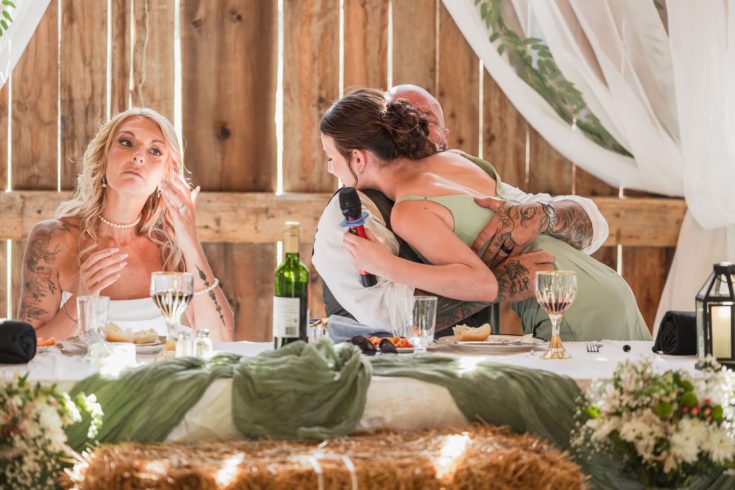 Wedding guests embrace at a rustic barn reception table adorned with greenery, wine bottles, and floral arrangements.