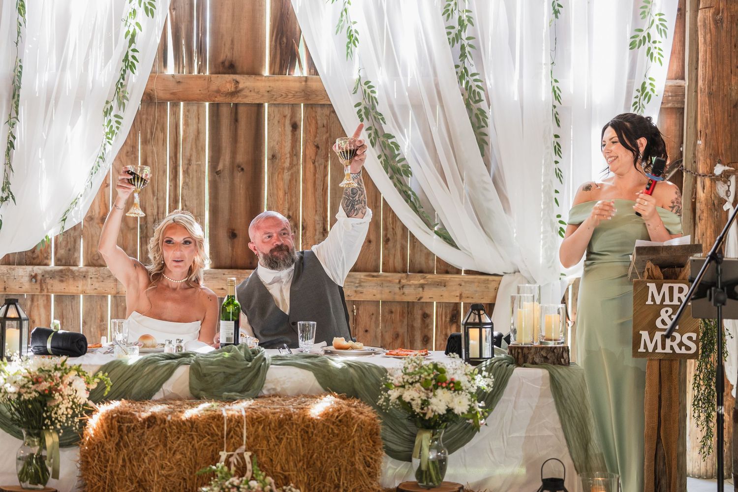 Bride and groom raise glasses during wedding toast in rustic barn venue decorated with white drapes and greenery.