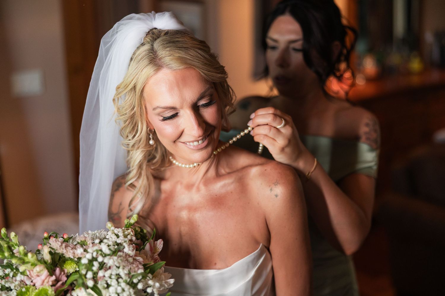Bridesmaid helping blonde bride put on pearl necklace while holding bouquet on wedding day.