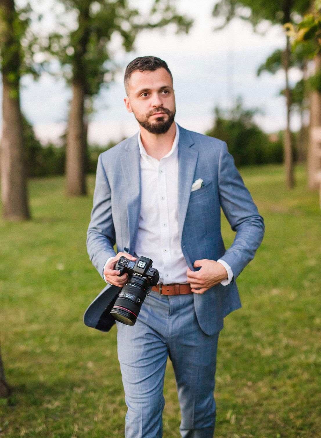 Wedding photographer in light blue suit adjusts camera settings in outdoor park setting.