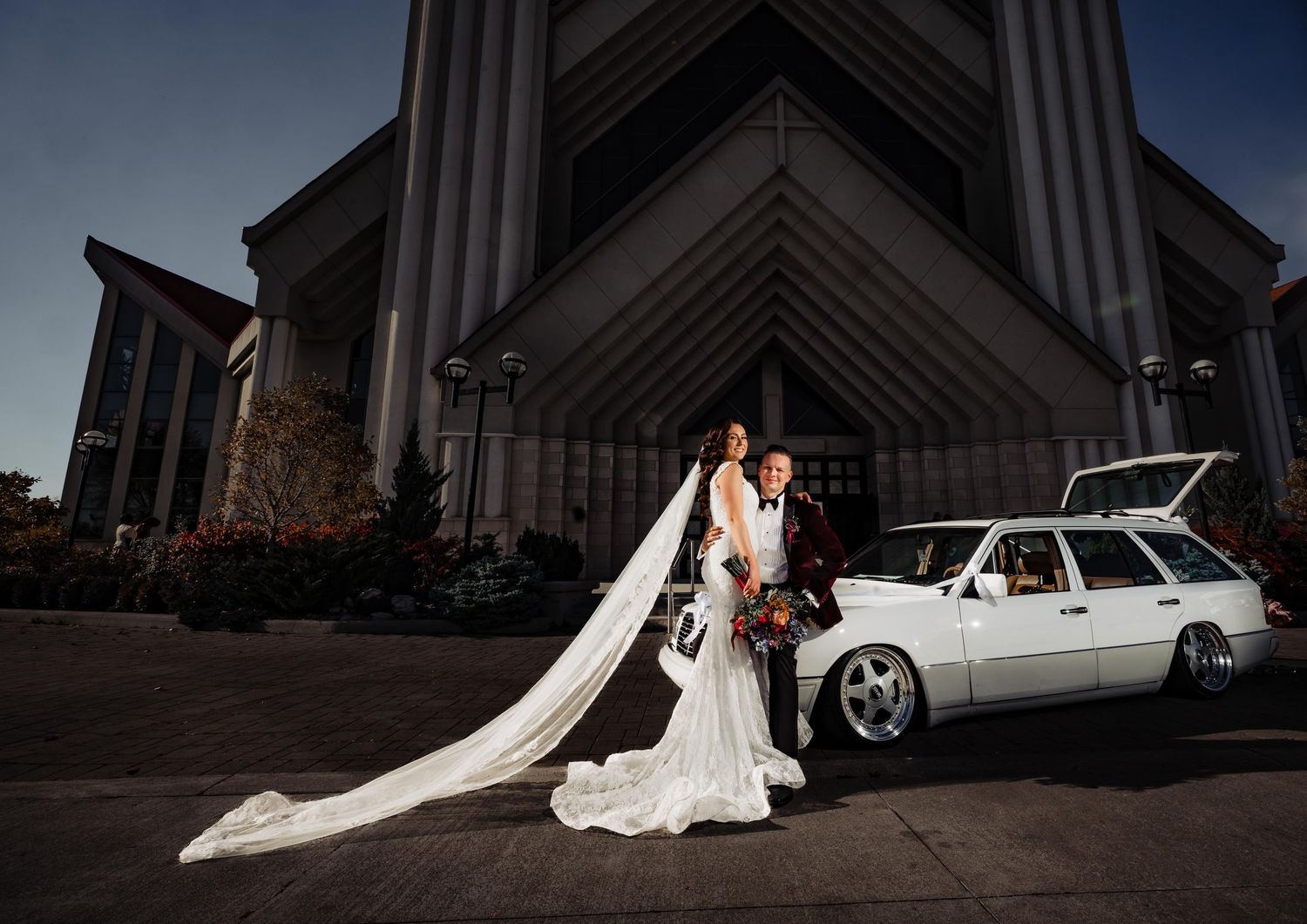 Wedding party poses outside modern angular church building with white car.