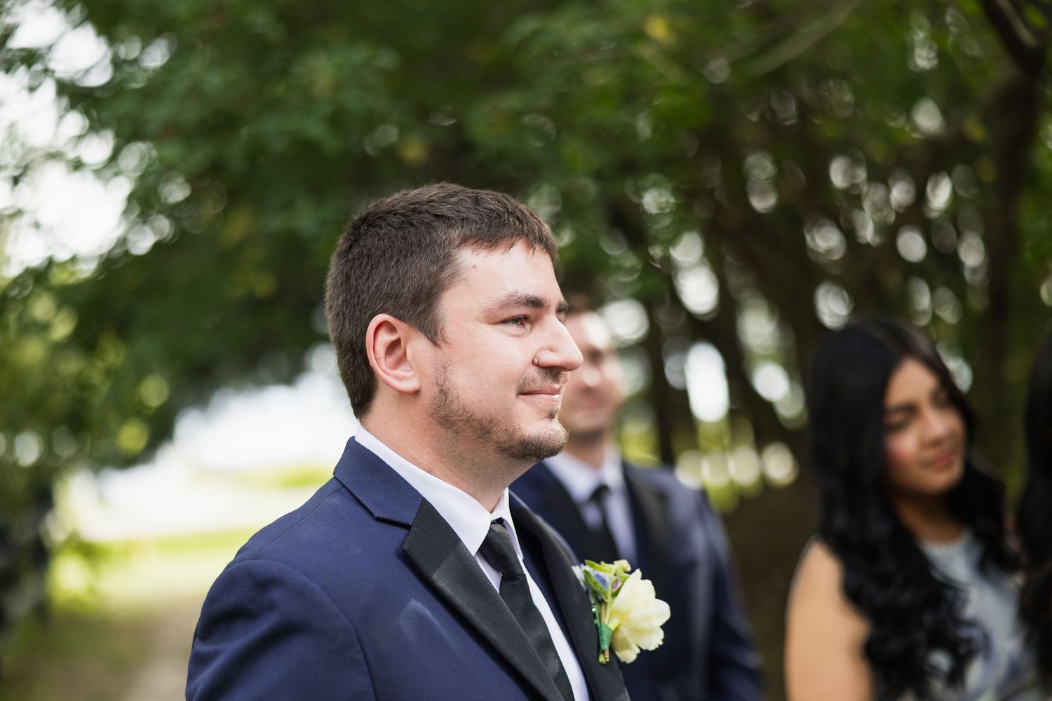 Smiling groom in navy suit with white boutonniere stands outdoors among trees at wedding.