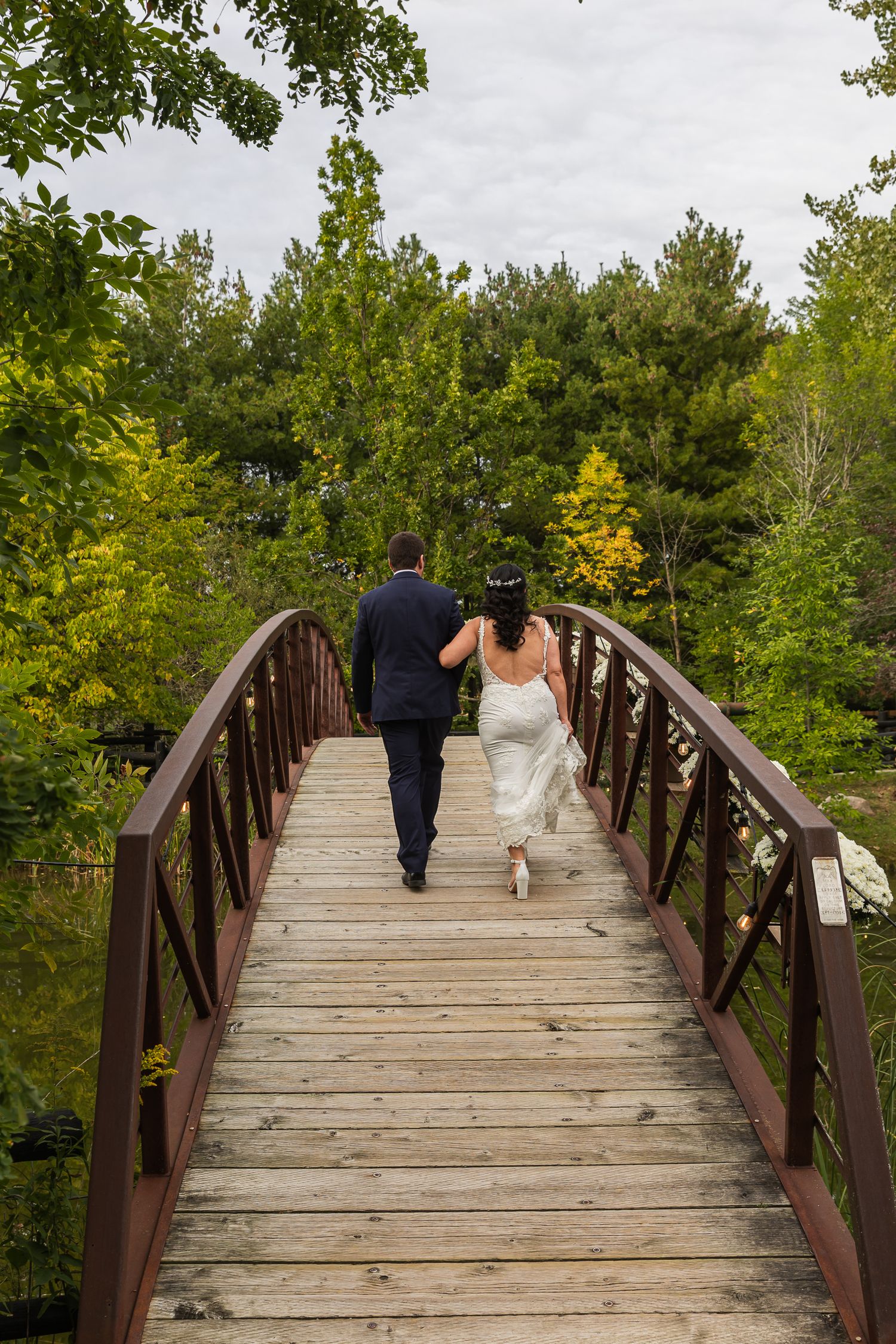 Bride and groom walking hand in hand across a wooden bridge surrounded by lush green trees on their wedding day.