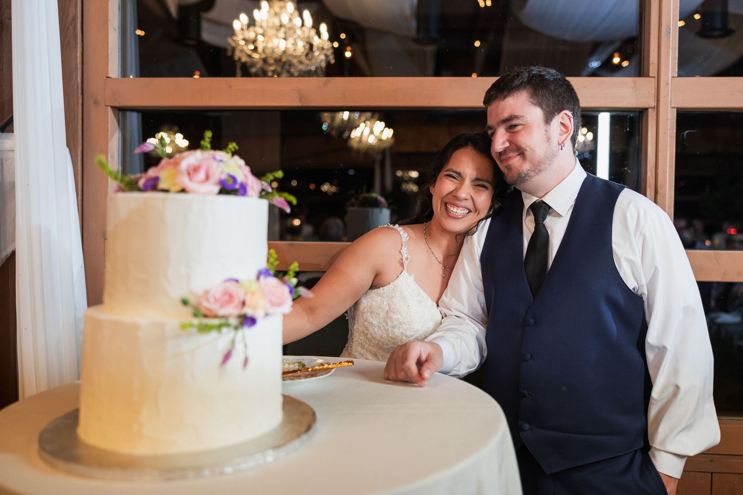 Smiling bride and groom pose together beside their two-tier white wedding cake adorned with colorful flowers.