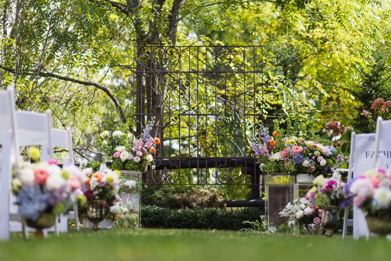 Elegant outdoor wedding ceremony setup with white chairs, colorful floral arrangements, and a decorative iron gate backdrop.