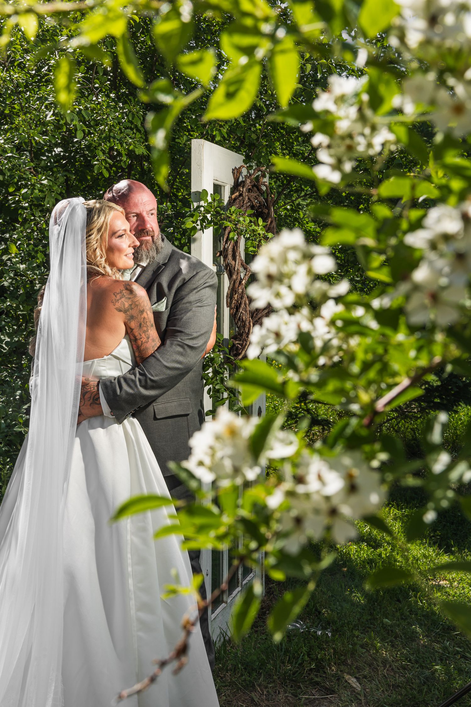 Bride and groom embrace under a white arch surrounded by lush greenery and white blooms on their wedding day.