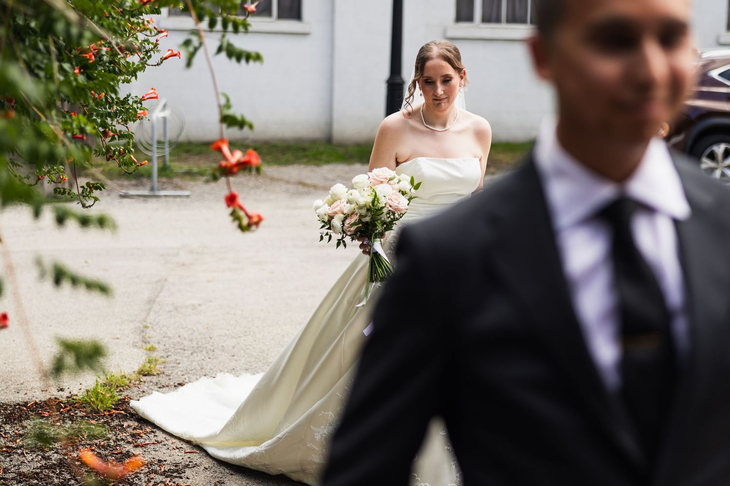 Bride in white gown holding bouquet approaches groom in dark suit during outdoor wedding ceremony.