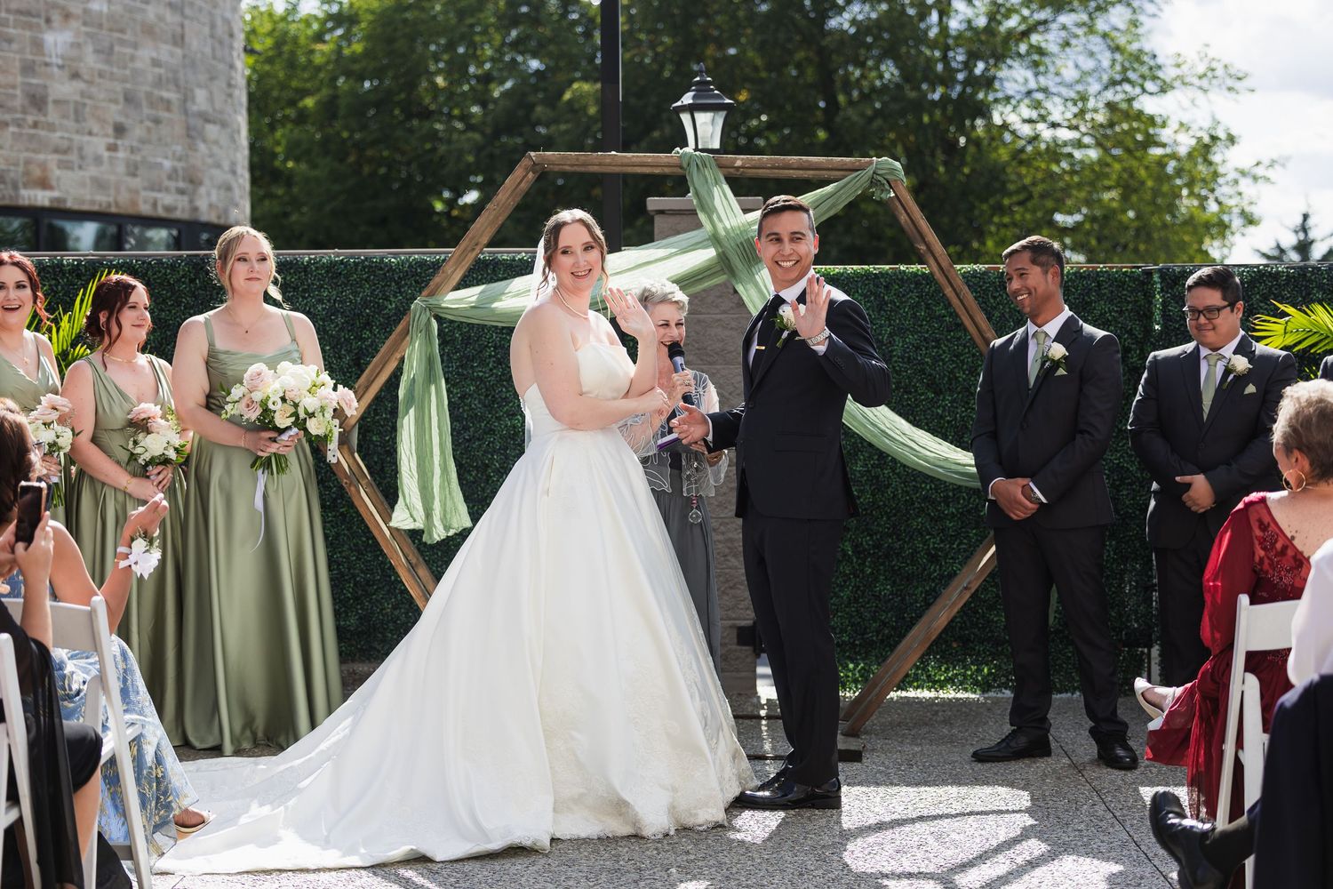 Bride and groom celebrate at outdoor wedding ceremony with bridal party beside hexagonal wooden arch.