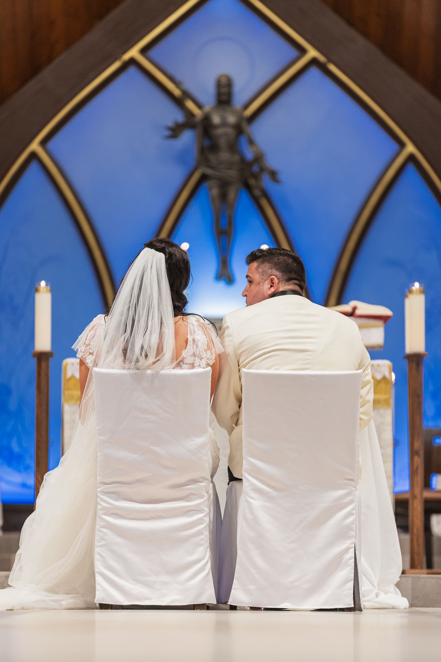 Bride and groom seated at the altar during their church wedding ceremony, facing a blue stained glass window.