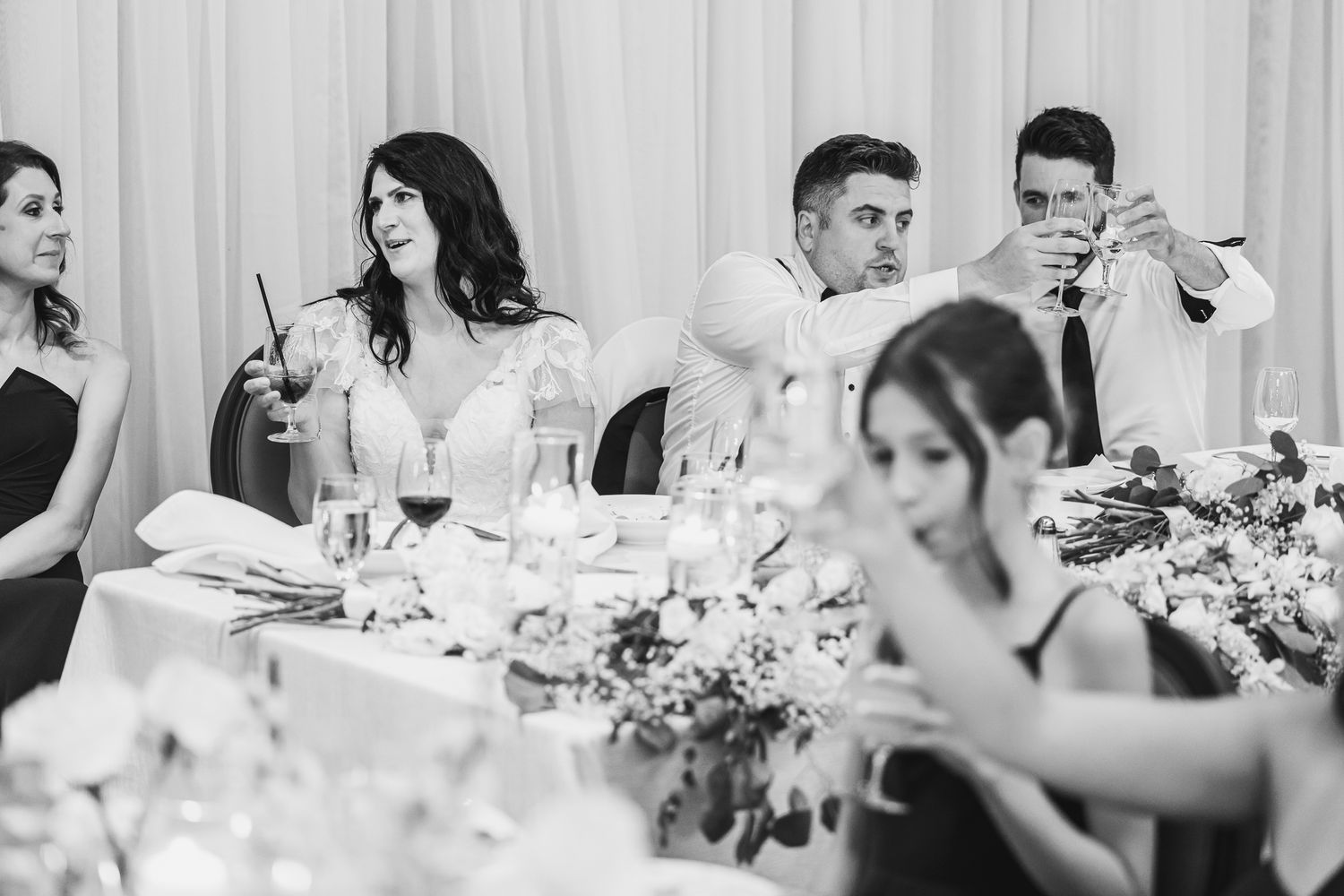 Black and white wedding reception photo of bride and groom seated at head table with guests celebrating in background.