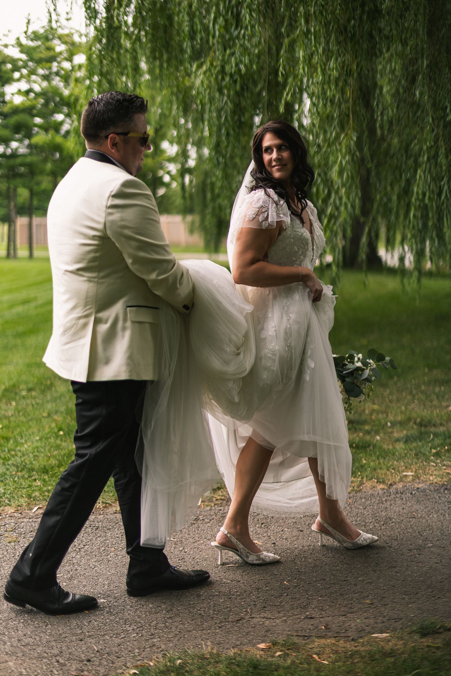 Groom carries bride's wedding dress train as she smiles walking along a garden path under willow trees.