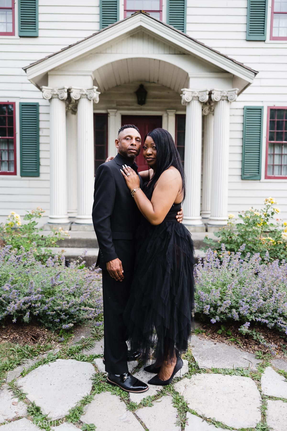 A couple in formal black attire poses together in front of an elegant white house with colonial style columns and a garden.