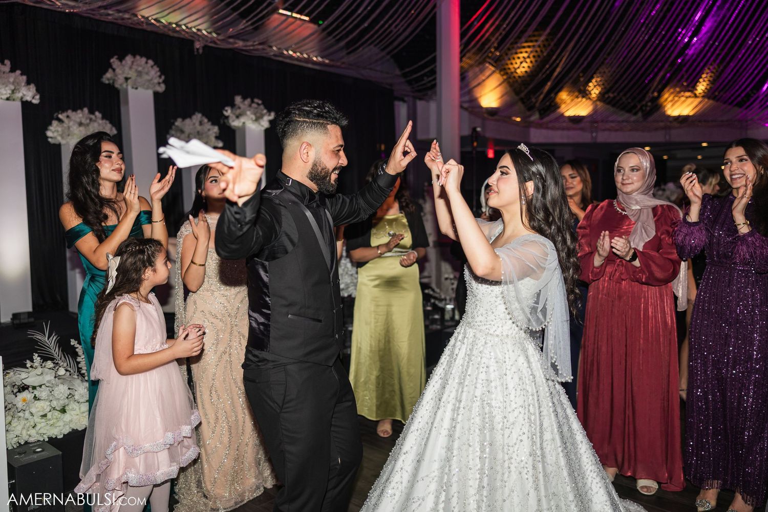 Wedding guests dance and celebrate on the dance floor at an indoor reception with colorful lighting and decorative columns.